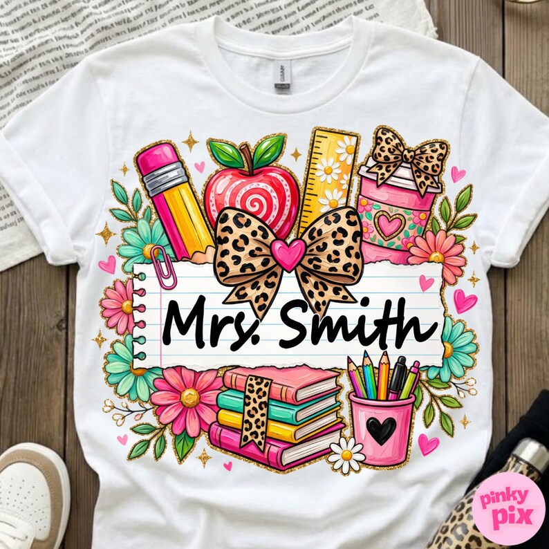 May include: White t-shirt featuring a colorful teacher-themed design. The design includes a pencil, apple, ruler, coffee cup, books, and pencils. The text "Mrs. Smith" is written on a lined paper. The design is surrounded by flowers and hearts.