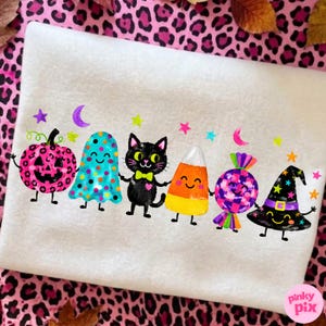 May include: A white sweatshirt featuring a colorful graphic print of a pink leopard-print pumpkin, a blue polka-dot ghost, a black cat with a yellow bow tie, a candy corn, a purple checkered candy, and a black witch hat, all with faces.