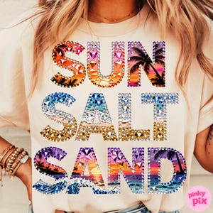 May include: Cream-colored t-shirt with the words "SUN SALT SAND" in a colorful, bejeweled design. The letters feature sunset and ocean scenes with palm trees. The shirt is a casual, beach-themed apparel item.