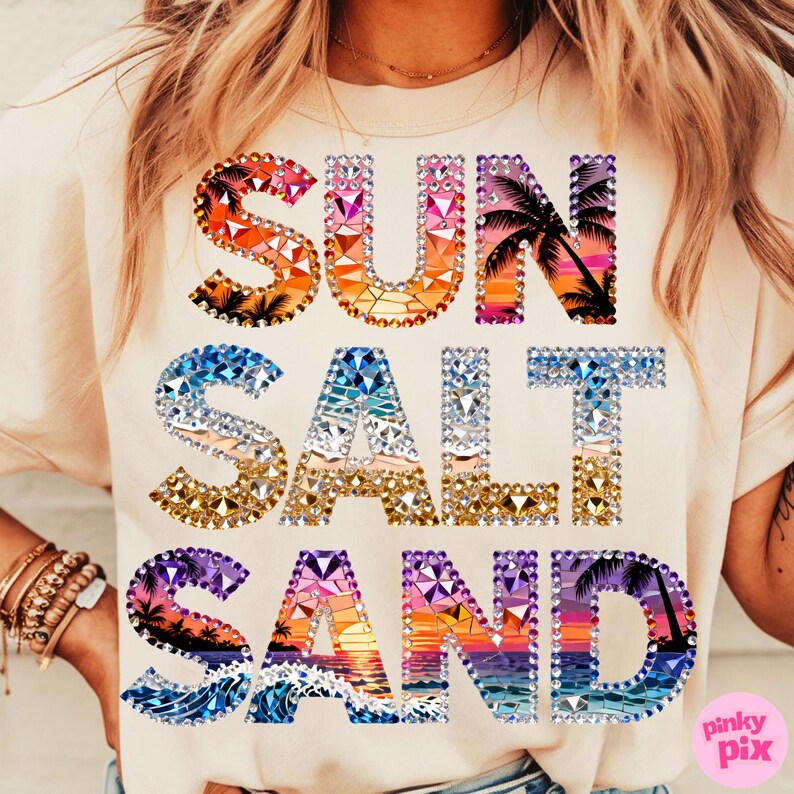 May include: Cream-colored t-shirt with the words "SUN SALT SAND" in a colorful, bejeweled design. The letters feature sunset and ocean scenes with palm trees. The shirt is a casual, beach-themed apparel item.