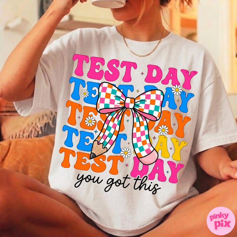 May include: White t-shirt with the words "TEST DAY" in colorful, retro-style lettering. A large bow with a checkered pattern and a pencil graphic are in the center. The phrase "you got this" is at the bottom.