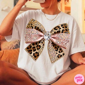 May include: White t-shirt featuring a large, embellished bow. The bow has a pink center, leopard print wings, and is adorned with rhinestones and gold accents. A large, clear gem is in the center of the bow.