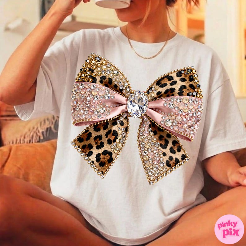 May include: White t-shirt featuring a large, embellished bow. The bow has a pink center, leopard print wings, and is adorned with rhinestones and gold accents. A large, clear gem is in the center of the bow.