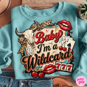 Może przedstawiać: Jasnoniebieska bluza z nadrukiem graficznym. Projekt zawiera czaszkę byka, karty do gry, kości, wiśnie oraz napis "Baby I'm a Wildcard" w kolorze czerwonym i czarnym. Widnieje r&oacute;wnież liczba 777.
