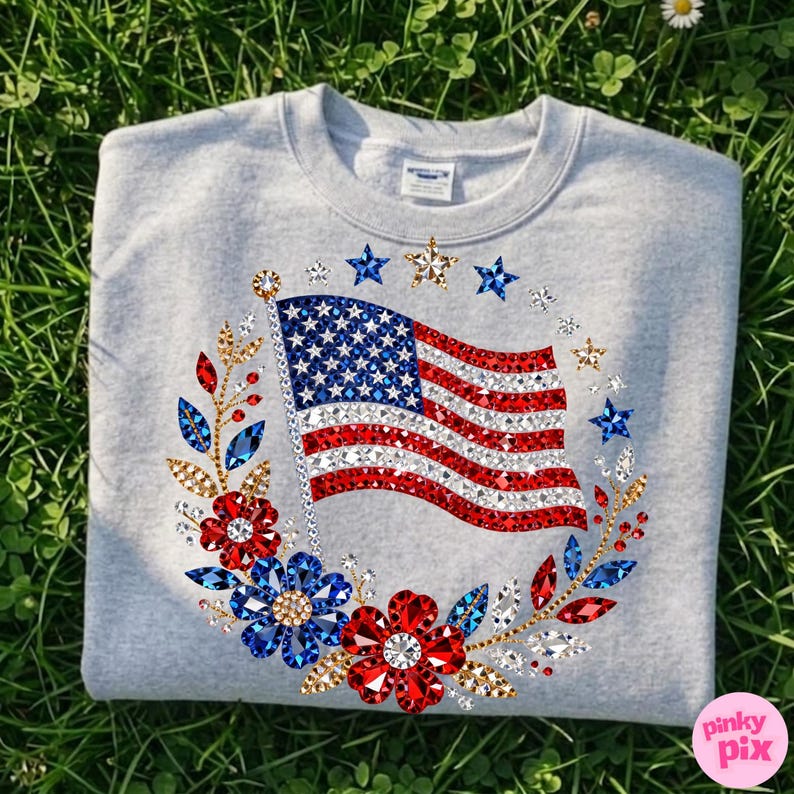 May include: A light gray sweatshirt featuring a sparkling American flag design. The flag is surrounded by a wreath of red, blue, and gold floral and star embellishments. The design is centered on the front of the sweatshirt.