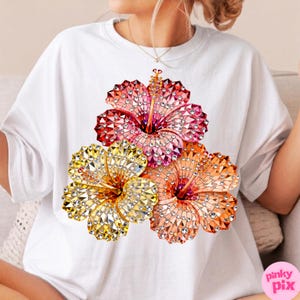 May include: White t-shirt featuring a vibrant floral design. The design showcases three hibiscus flowers in shades of red, pink, orange, and yellow, embellished with sparkling details. The flowers are arranged in a cluster, creating a visually appealing graphic.