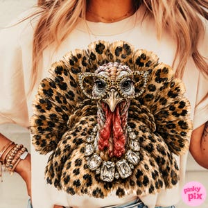 May include: A light beige t-shirt featuring a graphic print of a turkey with a leopard print pattern on its feathers. The turkey wears leopard print glasses and a large, clear crystal statement necklace around its neck.