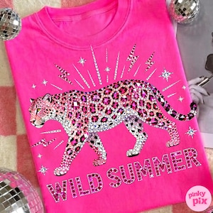 May include: A bright pink sweatshirt featuring a bedazzled leopard design with pink and clear rhinestones. The words "WILD SUMMER" are spelled out in pink rhinestones below the leopard. The design includes lightning bolts and stars.