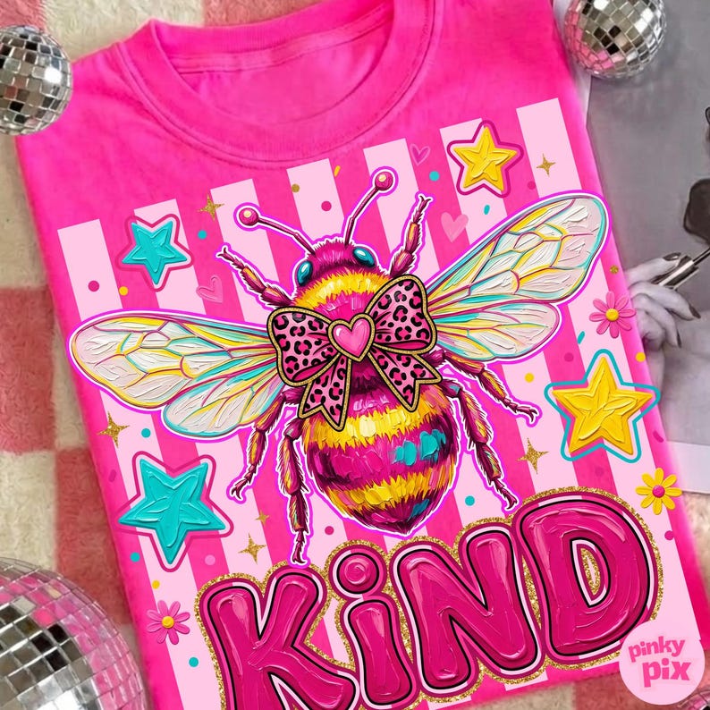 May include: A pink t-shirt featuring a large, colorful illustration of a bee with a leopard print bow and the word "KIND" in bubble letters. The design includes stars and stripes.
