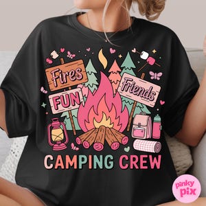 May include: Black t-shirt with a camping-themed graphic. The design includes a campfire, trees, signs that say "Fires", "Fun", and "Friends", and the words "Camping Crew". Pink, orange, and green colors are used.