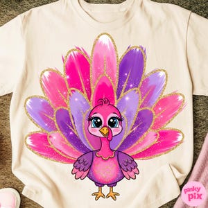 May include: A cream-colored t-shirt featuring a cartoon turkey with a vibrant, colorful design. The turkey has a pink body, purple wings, and a fanned tail in shades of pink and purple, accented with gold glitter. The turkey has big blue eyes.