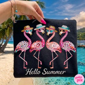 May include: Black tote bag featuring four pink flamingo illustrations, each adorned with hats and sunglasses. The flamingos are embellished with sparkling gems. The words "Hello Summer" are printed in white script. The background shows a tropical beach scene.