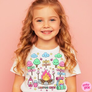 May include: White t-shirt with a colorful camping-themed graphic. The design includes a campfire, trees, clouds, and the words "Camping Crew." The shirt features a cute, cartoonish style with pastel colors and is suitable for children.