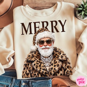 以下が含まれることがあります： クリーム色のクルーネックのスウェットシャツ。ブラウンの文字で「MERRY」と書かれ、その下にヒョウ柄の帽子とコート、サングラスを着用し、星のペンダントが付いた大きなクリスタルのステートメントネックレスを身に着けたサンタクロースのグラフィックがプリントされています。