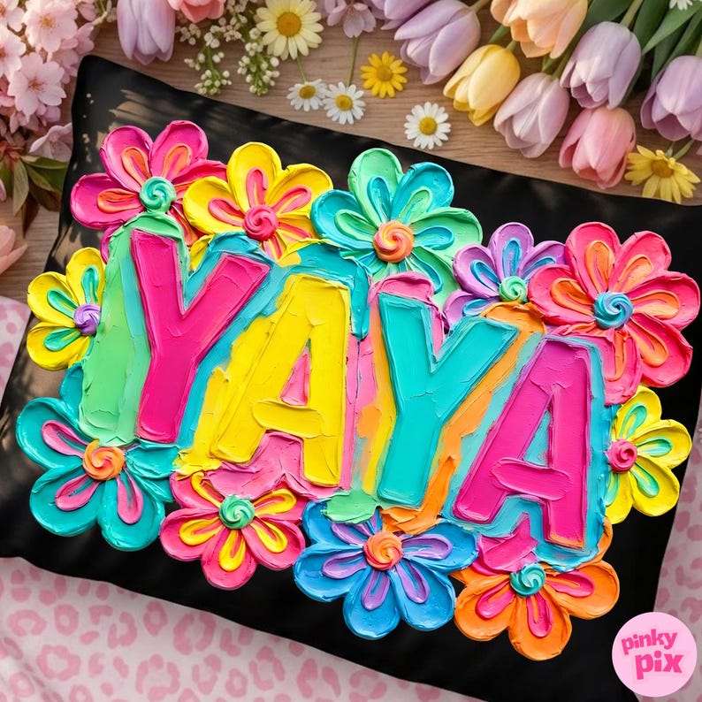 May include: Black decorative pillow with the word "YAYA" in bright, textured paint, surrounded by colorful, 3D flowers. The flowers are in shades of pink, yellow, blue, and orange. The pillow is on a pink and white patterned surface.