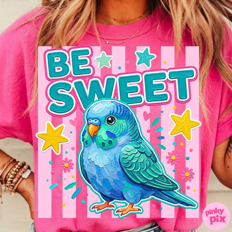 May include: Pink t-shirt with a graphic featuring a blue parakeet and the words "BE SWEET" in teal. The design includes stars, flowers, and confetti on a pink striped background.