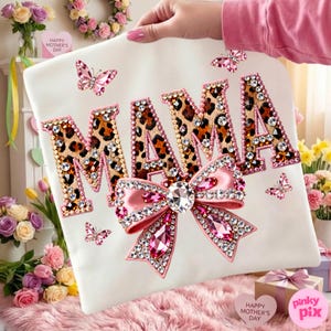 May include: A white t-shirt with the word "MAMA" in leopard print letters, outlined in pink and embellished with rhinestones. A large pink bow with a central rhinestone and smaller pink rhinestones is below the word. Pink butterfly accents complete the design.