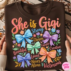 K&ouml;nnte beinhalten: Ein braunes Sweatshirt mit dem Text "She is Gigi" in Rosa und Gelb. Das Design umfasst bunte Schleifen und florale Akzente mit W&ouml;rtern wie "Strong", "Beautiful" und "Victorious".