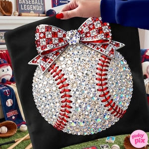 May include: A baseball-themed decorative pillow featuring a baseball design covered in sparkling rhinestones. The baseball has red stitching and is topped with a red and white checkered bow with a large rhinestone center. The pillow is on a black background.