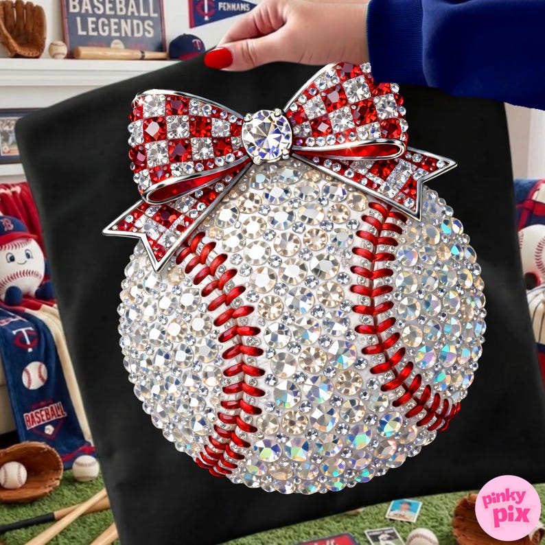 May include: A baseball-themed decorative pillow featuring a baseball design covered in sparkling rhinestones. The baseball has red stitching and is topped with a red and white checkered bow with a large rhinestone center. The pillow is on a black background.
