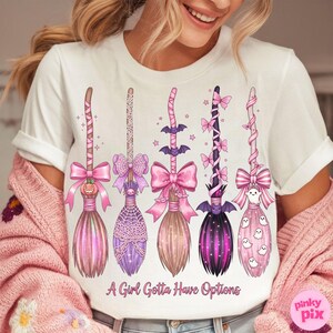 May include: White t-shirt featuring five whimsical broom designs in pink, purple, and brown hues. Each broom is adorned with bows, bats, and other Halloween-themed embellishments. The text "A Girl Gotta Have Options" is printed at the bottom.