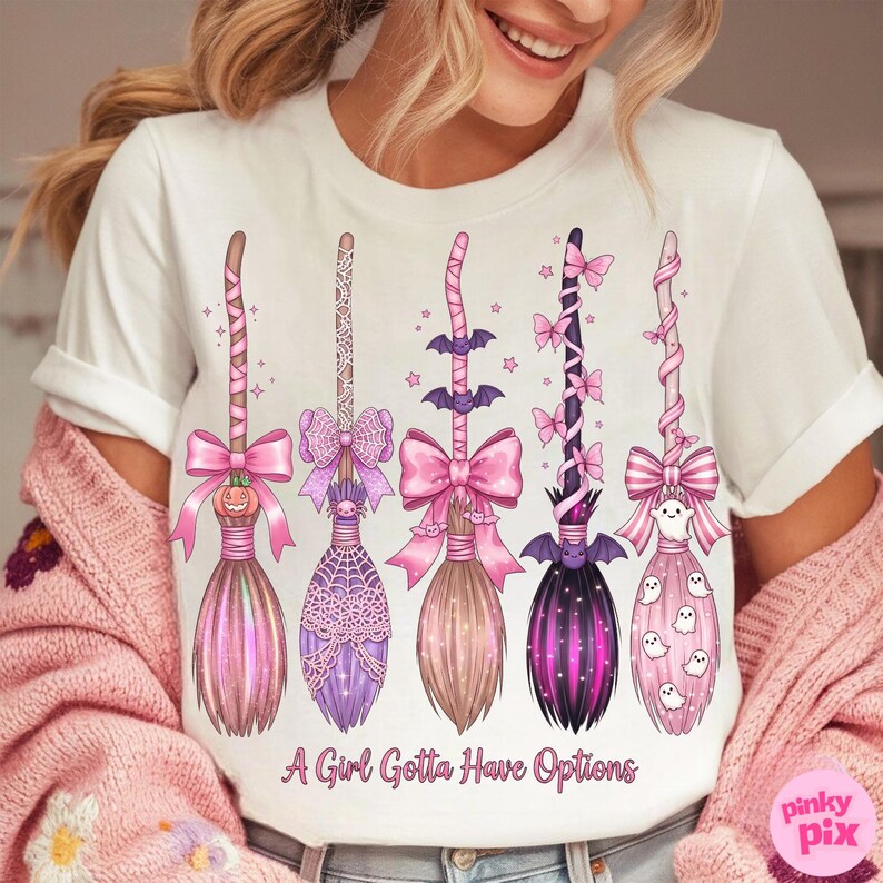 May include: White t-shirt featuring five whimsical broom designs in pink, purple, and brown hues. Each broom is adorned with bows, bats, and other Halloween-themed embellishments. The text "A Girl Gotta Have Options" is printed at the bottom.
