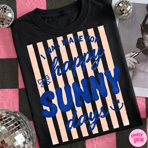 May include: Black t-shirt with the text "I WAS MADE FOR happy SUNNY days" in blue and peach. The design includes a bow and a smiling face. The shirt is a crew neck.