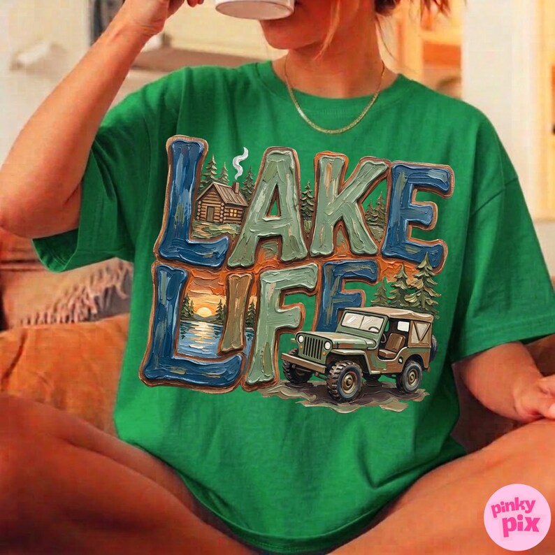 May include: Green t-shirt with the words "LAKE LIFE" in a textured, colorful design. The design includes a cabin, a sunset over a lake, and a green jeep. The shirt is a comfortable, casual style.
