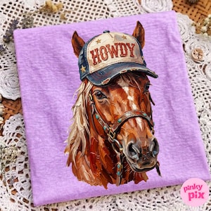 May include: A lilac-colored t-shirt featuring a detailed illustration of a horse wearing a baseball cap. The cap is navy blue and tan with the word "HOWDY" and star accents. The horse has a brown coat and a leather bridle.