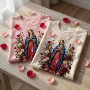 May include: Two crew neck t-shirts, one pink and one cream, each featuring a religious illustration of a woman surrounded by roses. The woman is wearing a blue cloak and a pink dress. Scattered rose petals add a decorative touch.