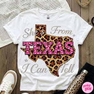 May include: White t-shirt with a Texas-shaped design in leopard print and pink text that reads "TEXAS". The shirt also features the text "She's From I Can Tell" in a sparkling font. A casual, stylish tee.