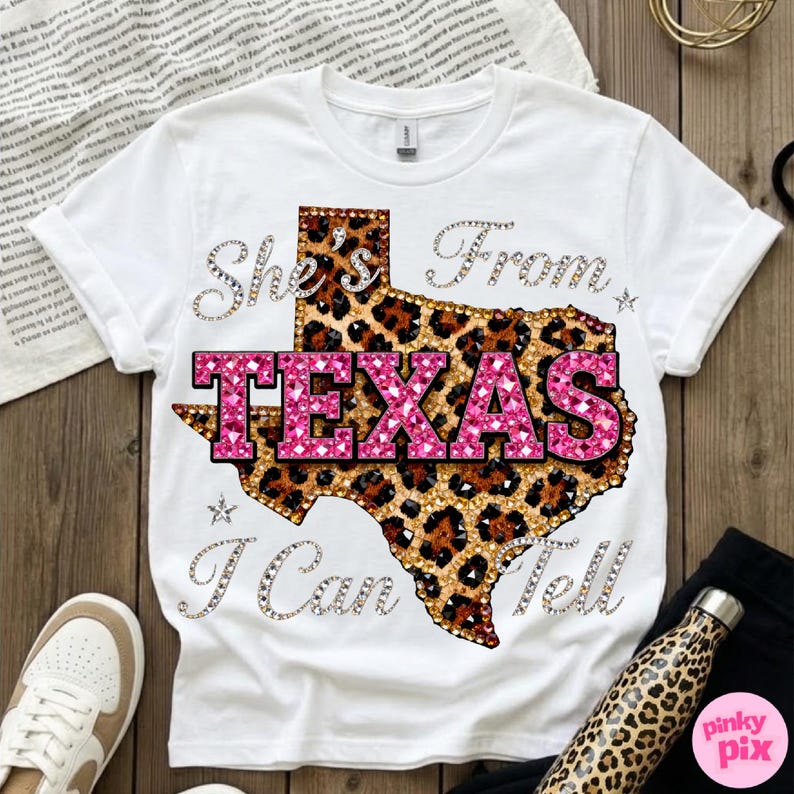 May include: White t-shirt with a Texas-shaped design in leopard print and pink text that reads "TEXAS". The shirt also features the text "She's From I Can Tell" in a sparkling font. A casual, stylish tee.