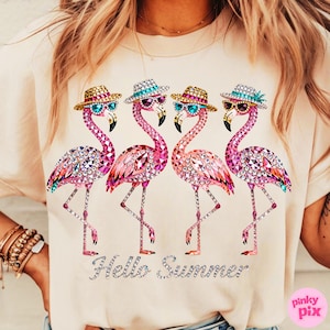 May include: A light beige t-shirt featuring four pink flamingo illustrations. Each flamingo wears sunglasses and a hat. The phrase "Hello Summer" is written in silver glitter below the flamingos. The shirt is a casual summer wear.