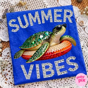 May include: Blue t-shirt with the words "SUMMER VIBES" in silver glitter. A bejeweled turtle wearing sunglasses floats on an inflatable ring. The shirt has a Pinky Pix logo.