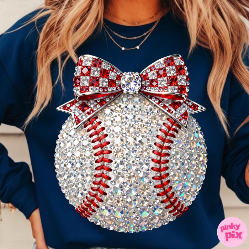 May include: Navy blue sweatshirt featuring a baseball design. The baseball is covered in sparkling crystals with red stitching. A red and white checkered bow with a large crystal center adorns the top of the baseball.