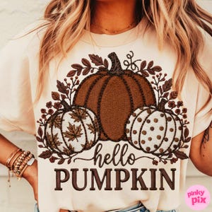 Puede incluir: Camiseta color crema con un diseño de calabaza marrón y dos calabazas más pequeñas a cada lado. Una tiene un patrón de hojas de arce y la otra lunares marrones. El texto "hello PUMPKIN" está debajo.