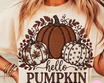 May include: A cream-coloured t-shirt with a brown pumpkin design. Two smaller pumpkins flank the central pumpkin, one with a maple leaf pattern, the other with brown polka dots. The text "hello PUMPKIN" is below.