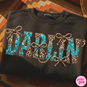 Pu&ograve; includere: Felpa nera con la scritta "DARLIN'" in turchese, stampa leopardata e dettagli in corda. Le lettere sono riempite di pietre turchesi e incorniciate da stampa leopardata. Fiocchi e stelle di corda aggiungono dettagli.