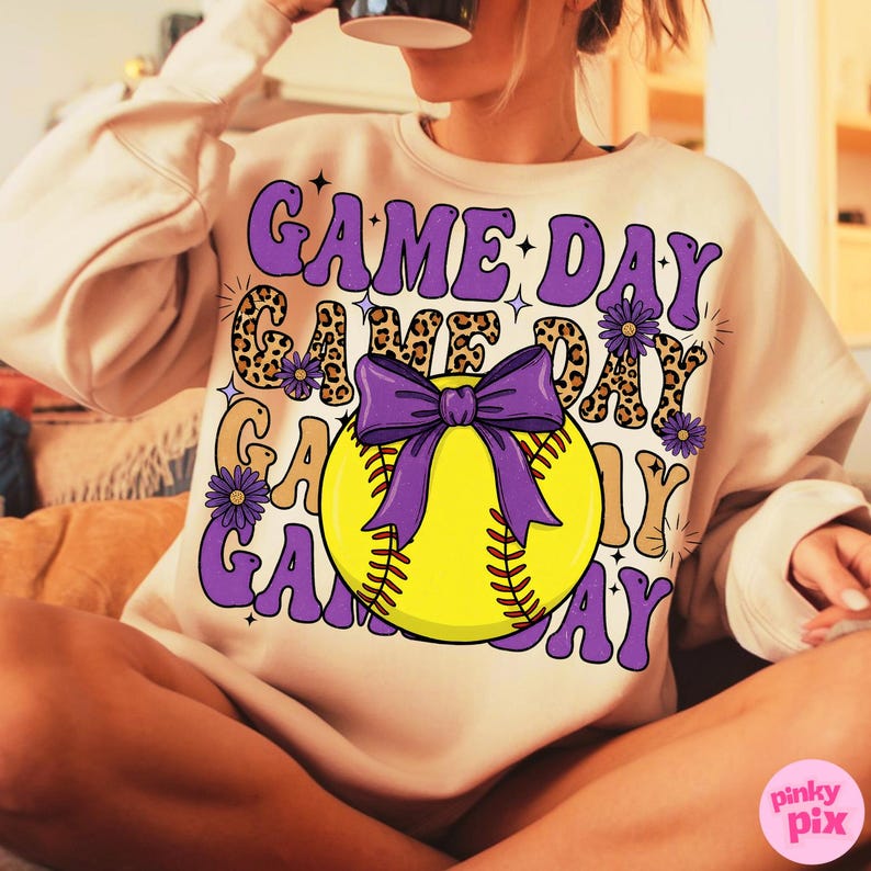 May include: A beige sweatshirt with a graphic design featuring the words "GAME DAY" in purple, leopard print, and yellow. A yellow softball with a purple bow is in the center. Purple flowers and stars complete the design. The sweatshirt is a casual sports-themed apparel.