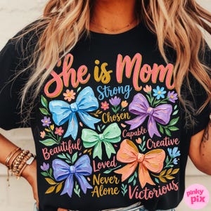 K&ouml;nnte beinhalten: Schwarzes T-Shirt mit den Worten "She is Mom" in rosa und gelber Schrift. Das Shirt zeigt bunte Schleifen und florale Akzente mit W&ouml;rtern wie "Strong", "Beautiful" und "Victorious".