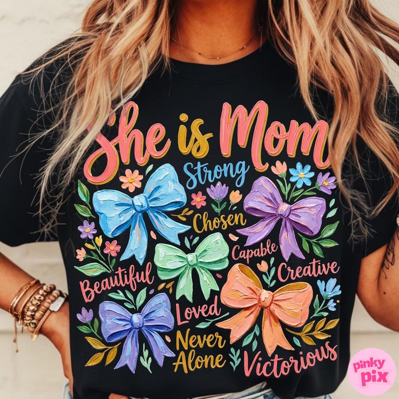 K&ouml;nnte beinhalten: Schwarzes T-Shirt mit den Worten "She is Mom" in rosa und gelber Schrift. Das Shirt zeigt bunte Schleifen und florale Akzente mit W&ouml;rtern wie "Strong", "Beautiful" und "Victorious".