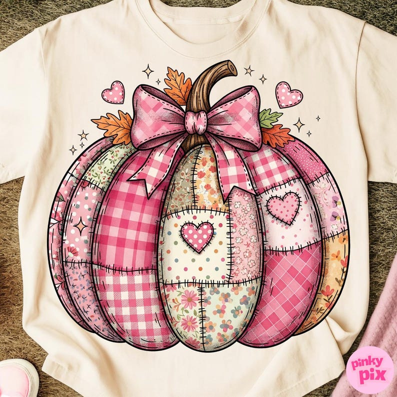 May include: Cream-colored t-shirt featuring a patchwork pumpkin design. The pumpkin is composed of various pink patterns, including gingham and floral prints, with a pink bow and heart accents. The design has a stitched appearance.