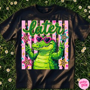May include: Black t-shirt featuring a green alligator wearing heart-shaped sunglasses and giving a peace sign. The word "later" is written in green script above the alligator. Pink and white striped background with star and confetti accents.