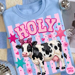May include: Light blue sweatshirt with a graphic of a cow wearing a leopard print bow and the word "HOLY" in pink. The cow has black and white spots and is surrounded by pink stars, hearts, and flowers.