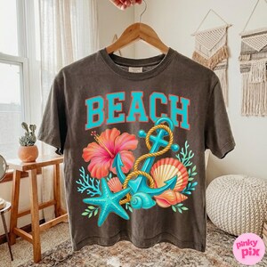 May include: A dark grey t-shirt featuring the word "BEACH" in turquoise lettering. The design includes a turquoise anchor, rope, a pink hibiscus flower, a blue starfish, and seashells.