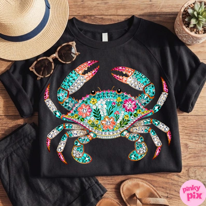 May include: Black t-shirt featuring a colorful crab design. The crab is decorated with turquoise, pink, and white gems, and floral accents. The design is centered on the shirt, with a straw hat and sunglasses in the background.