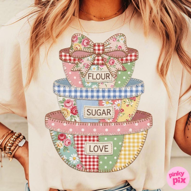 May include: Cream-colored t-shirt featuring a stack of patchwork mixing bowls with a bow at the top. The bowls are decorated with floral, gingham, and polka dot patterns. Text on the bowls reads "FLOUR", "SUGAR", and "LOVE".