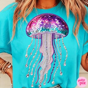 May include: A turquoise t-shirt featuring a sequined jellyfish design. The jellyfish has a purple, pink, and teal sequined head, with beaded and crystal-like strands hanging below. The design is centered on the shirt.
