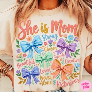 K&ouml;nnte beinhalten: Cremefarbenes T-Shirt mit dem Text "She is Mom" in Rosa und Gelb, umgeben von bunten Schleifen und floralen Akzenten. Das Shirt zeigt auch W&ouml;rter wie "Strong", "Beautiful" und "Victorious".
