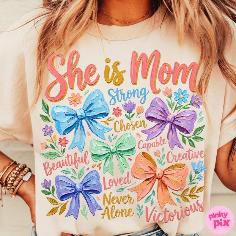 K&ouml;nnte beinhalten: Cremefarbenes T-Shirt mit dem Text "She is Mom" in Rosa und Gelb, umgeben von bunten Schleifen und floralen Akzenten. Das Shirt zeigt auch W&ouml;rter wie "Strong", "Beautiful" und "Victorious".
